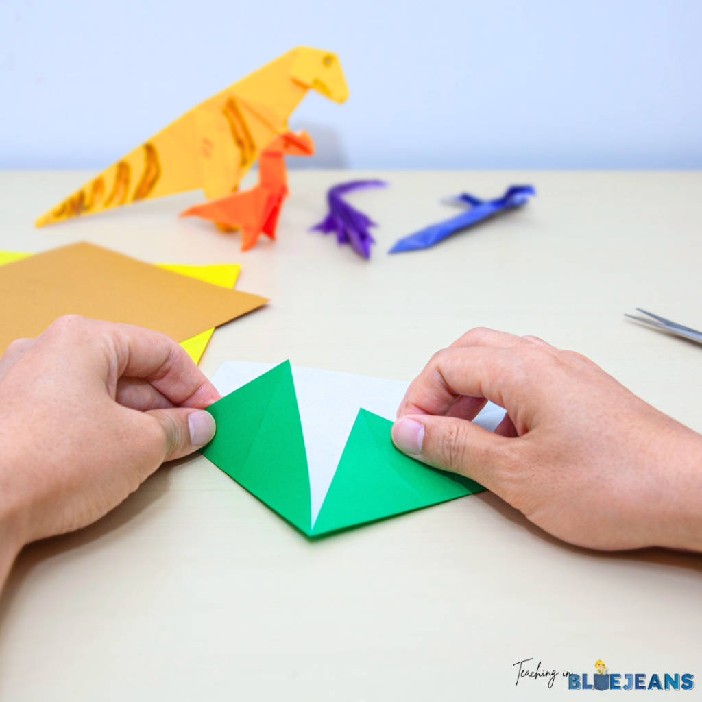 This image shows a child folding paper in order to make origami shapes. There are several dinosaur origami crafts in the background.