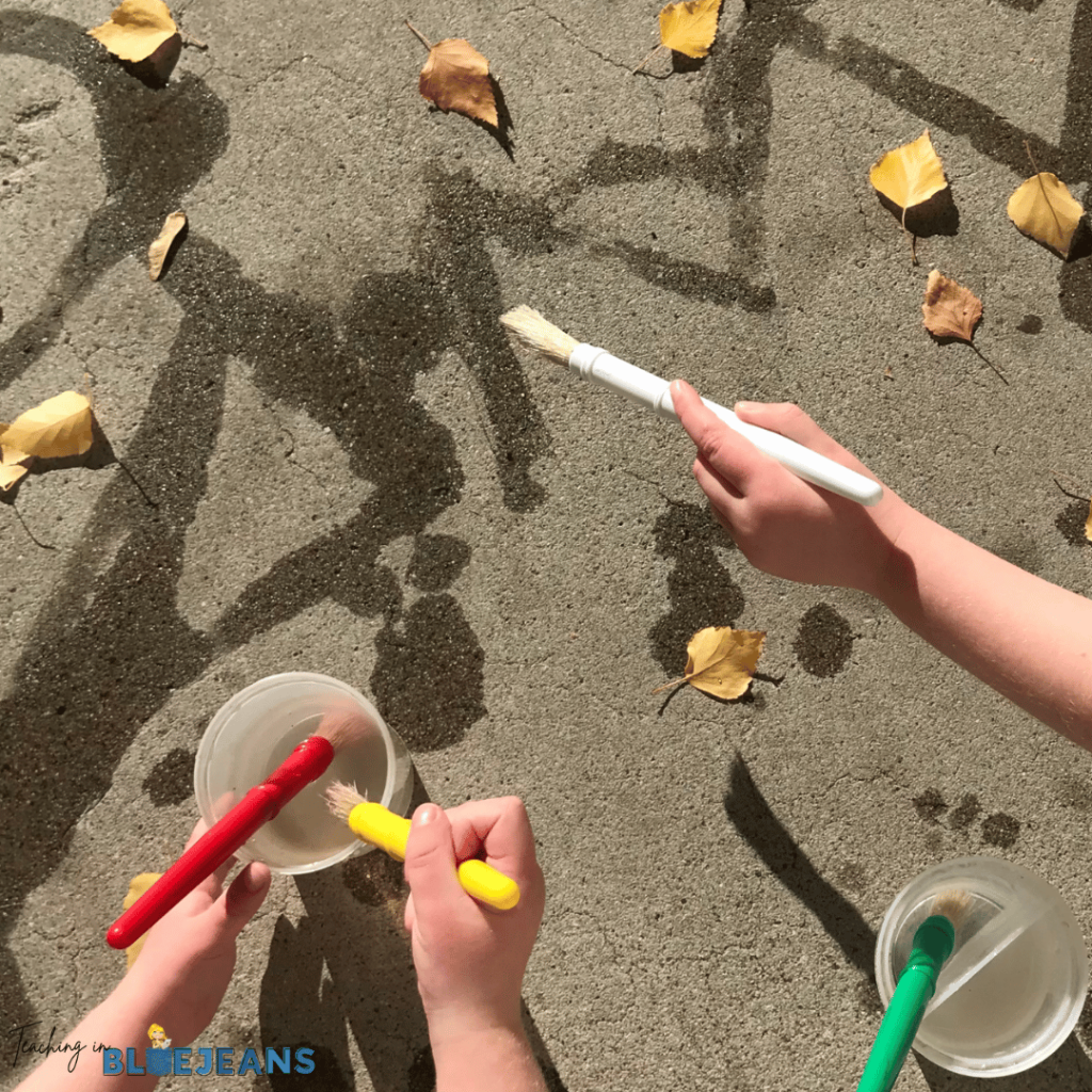 This photo shows young students using paintbrushes and water to paint shapes, lines and letters as a prewriting activity.